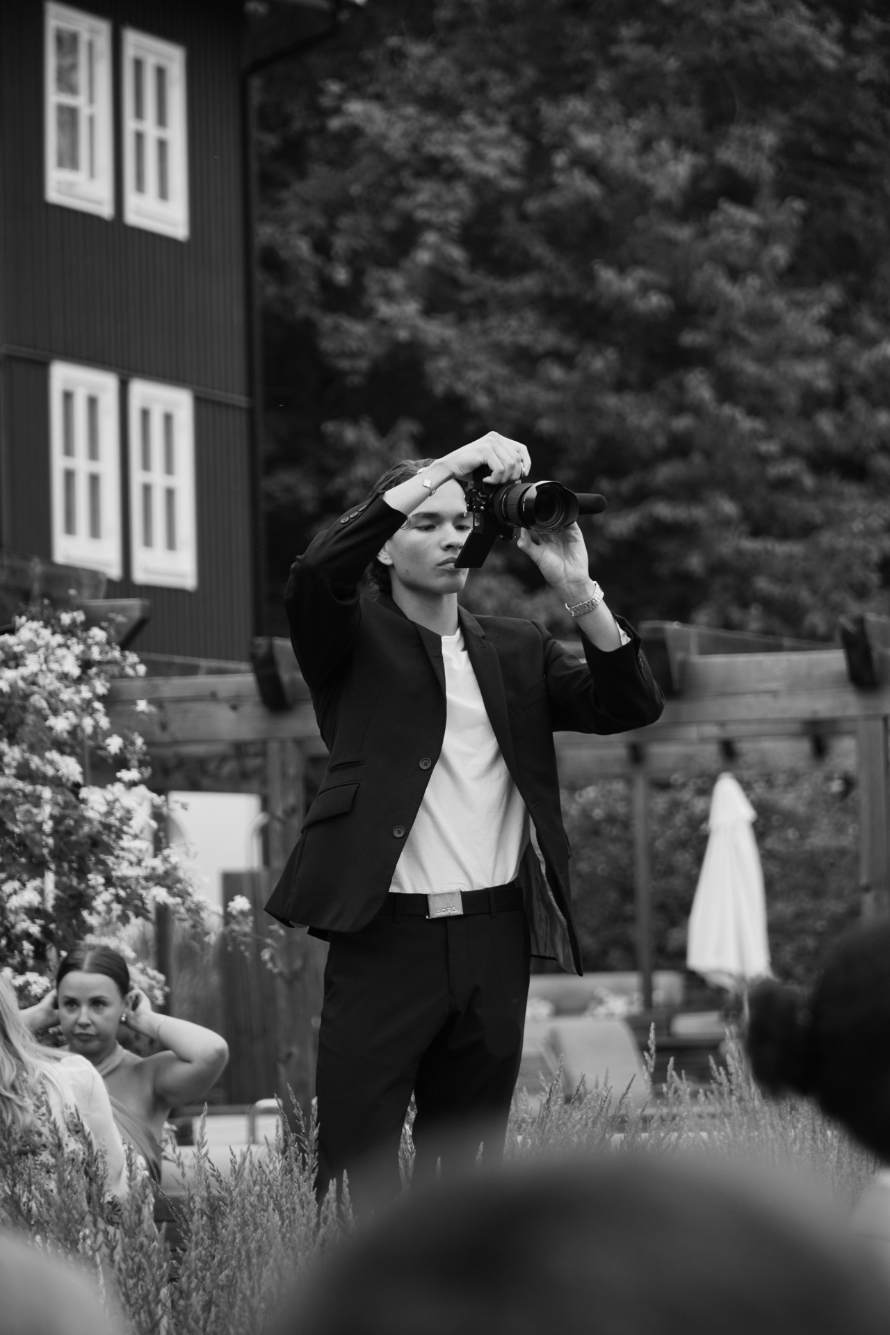 Svartvit bild utomhus av ung person i mörk kostym som håller en systemkamera med mikrofon bland gäster i en trädgård