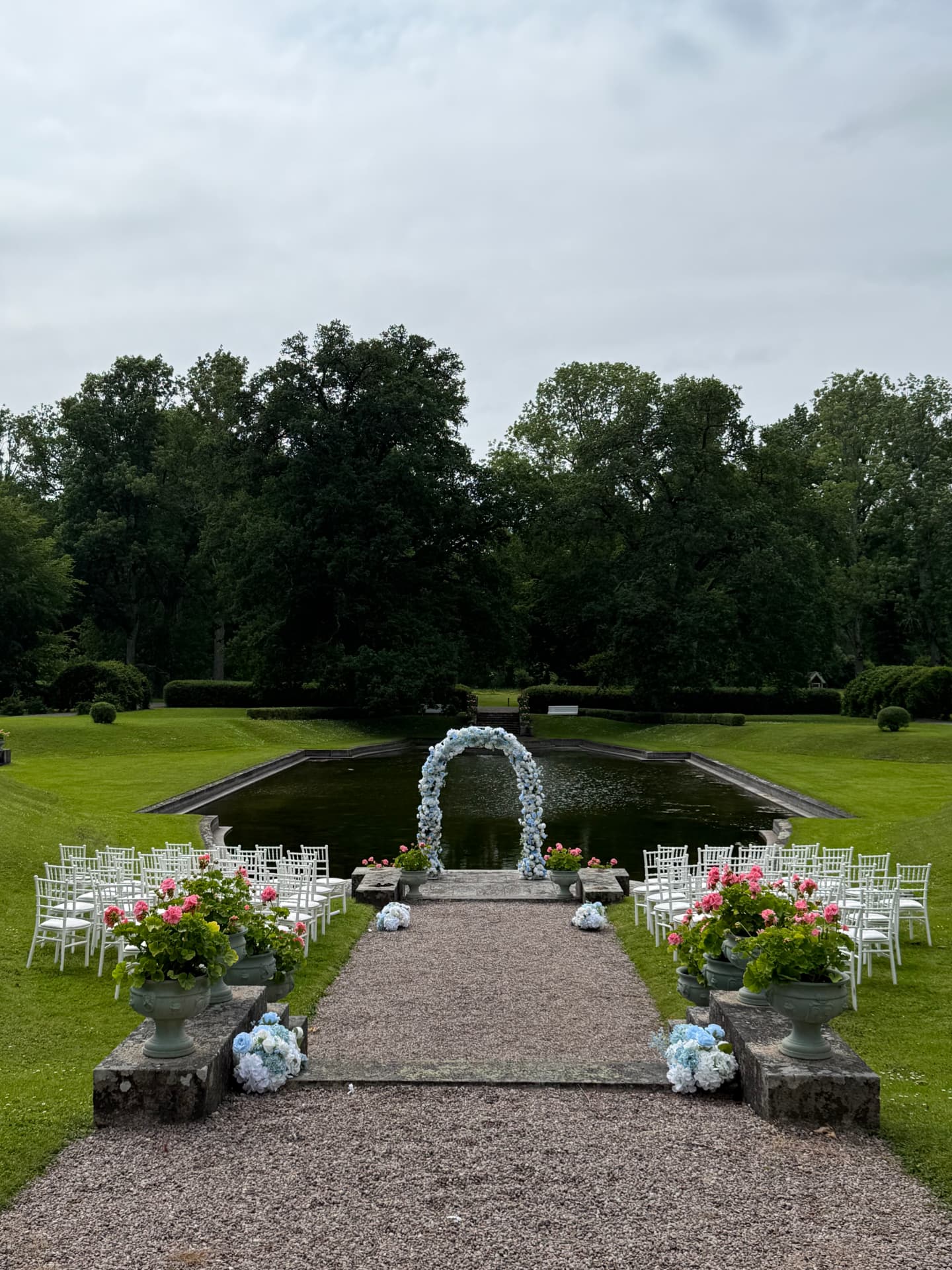 Utomhusceremoni med blomsterbåge, vita stolar och blomurnor vid dammen